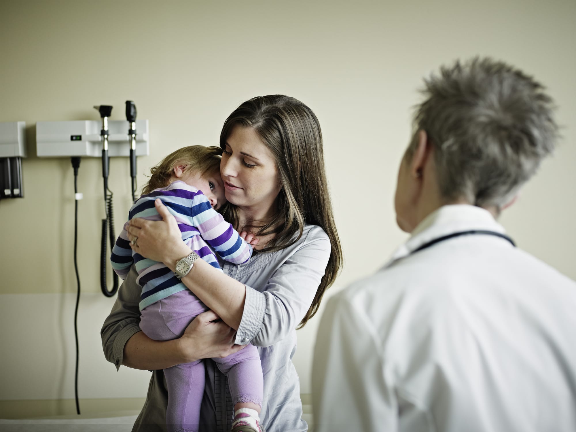 Mother embracing young daughter in exam room while in discussion with doctor